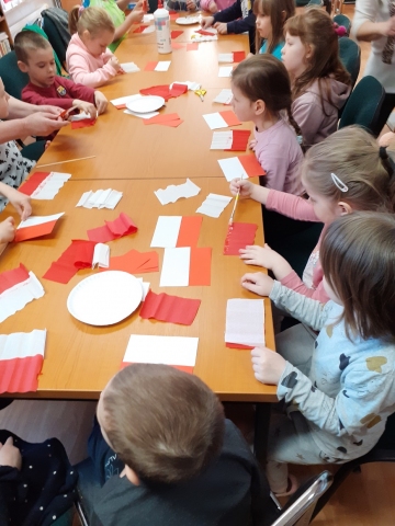 Zdjęcie prezentuje lekcję biblioteczną w Gminnej Bibliotece Publicznej w Wąwolnicy dotyczącą symboli narodowych Rzeczypospolitej Polskiej. Dzieci wykonują flagę. 