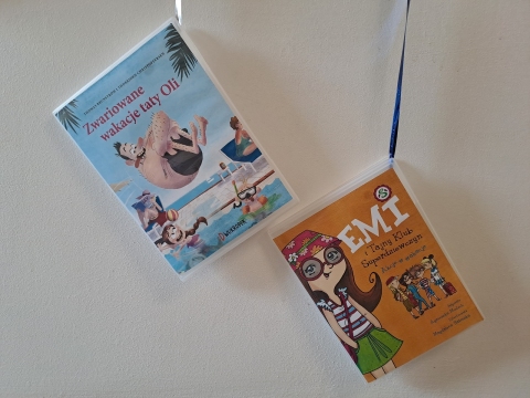 Zdjęcie prezentuje wystawę powieszoną na korytarzu przed biblioteką prezentującą różne okładki książek z wyrazem wakacje w tytule.