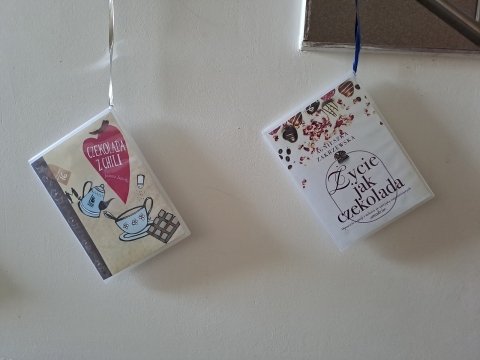 Zdjęcie prezentuje wystawę powieszoną na korytarzu przed biblioteką prezentującą różne okładki książek z wyrazem czekolada w tytule.