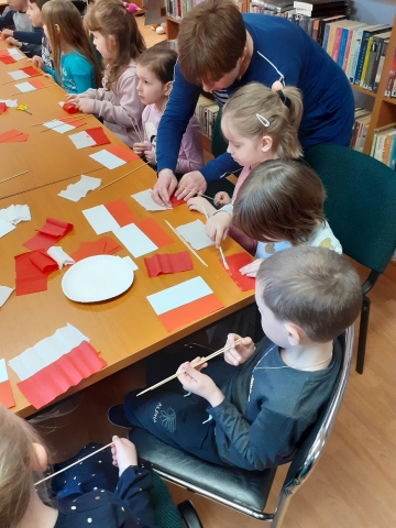 Zdjęcie prezentuje lekcję biblioteczną w Gminnej Bibliotece Publicznej w Wąwolnicy dotyczącą symboli narodowych Rzeczypospolitej Polskiej. Dzieci wykonują flagę. 
