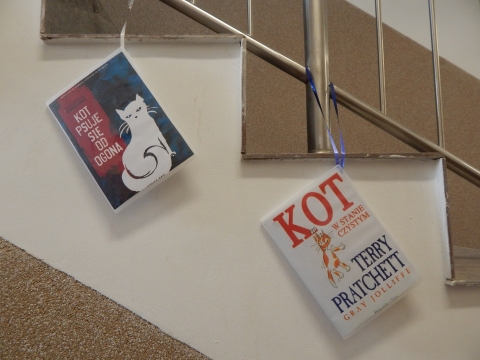 Zdjęcie prezentuje wystawę powieszoną na korytarzu przed biblioteką prezentującą różne okładki książek w których głównym bohaterem jest kot. 