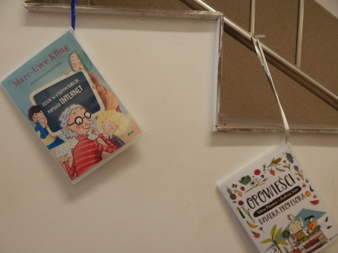 Zdjęcie prezentuje wystawę powieszoną na korytarzu przed biblioteką prezentującą różne okładki książek w których głównymi bohaterami są babcia i dziadek.