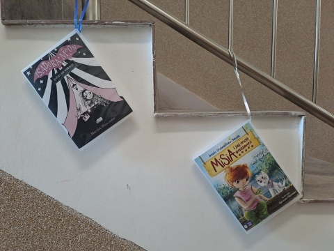 Zdjęcie prezentuje wystawę powieszoną na korytarzu przed biblioteką prezentującą różne okładki książek z wyrazem wakacje w tytule.