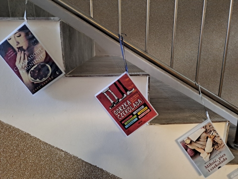 Zdjęcie prezentuje wystawę powieszoną na korytarzu przed biblioteką prezentującą różne okładki książek z wyrazem czekolada w tytule.