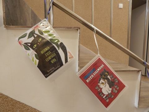 Zdjęcie prezentuje wystawę powieszoną na korytarzu przed biblioteką prezentującą różne okładki książek w których głównym bohaterem jest kot. 