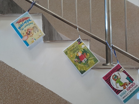 Zdjęcie prezentuje wystawę powieszoną na korytarzu przed biblioteką prezentującą różne okładki książek z wyrazem wakacje w tytule.