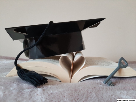 Zdjęcie przedstawia rozłożona książkę, w której środkowe kartki ułożone są w kształcie serca. Na książce położony jest biret absolwenta, a obok stoi klucz. 
