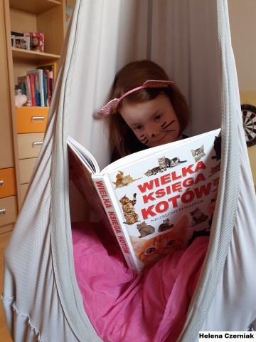 Zdjęcie przedstawia dziewczynkę siedząca w hamaku, przebraną za kota. Dziewczynka w ręce trzyma książkę pt. „Wielka Księga Kotów”. 