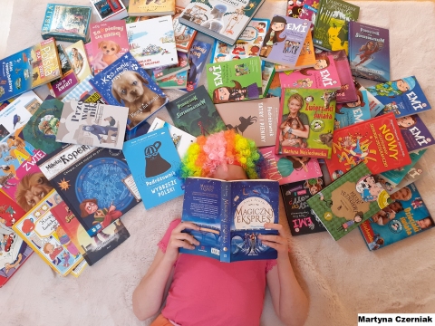 Zdjęcie przedstawia leżąca na dywanie dziewczynkę w kolorowej peruce na głowie. Obok niej ułożone są książki. 