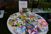 Zdjęcie prezentuje akcję bookcrossing podczas XVI Turniej Piłki Nożnej Samorządowców Powiatu Puławskiego im. Mariusza Próchniaka przygotowaną przez Powiatową Bibliotekę Publiczną w Puławach. Na zdjęciu ułożone na stoliku kolorowanki i biblioteczne zakładki oraz plakat promujący akcję. 
