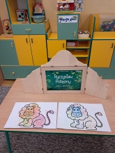 Zdjęcie prezentuje lekcję biblioteczną w Publicznym Przedszkolu Integracyjnym w Puławach. Na zdjęciu widać stół, na którym stoi drewniany teatrzyk Kamishibai z widoczną tytułową kartę bajki Sabiny Furmańskiej pt. „Ryczysław Potężny” oraz dwa pokolorowane rysunki lwa. 